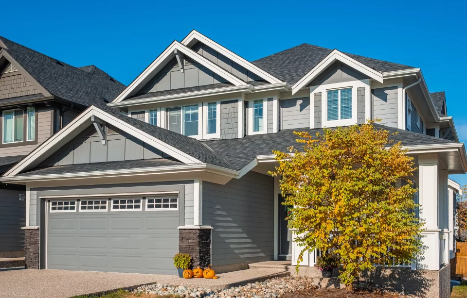 Gray suburban house in Tampa with a gabled roof, autumn tree, and pumpkins by the garage under a clear blue sky.